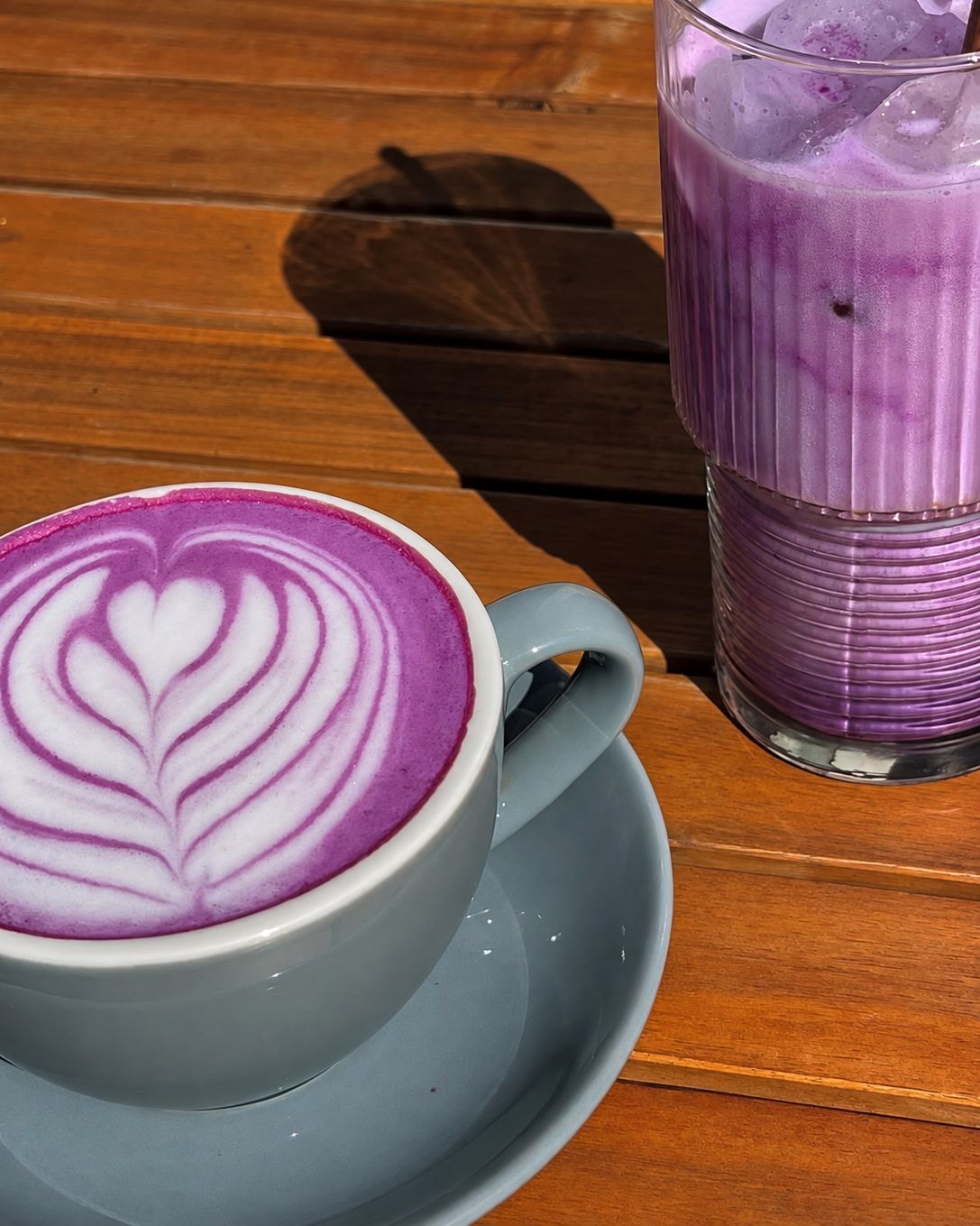 Purple ube latte with latte art and an iced ube latte on a wooden café table in the sun